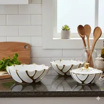 Trois saladiers design blanc et or disposés séparément avec accessoires de cuisine en fond.